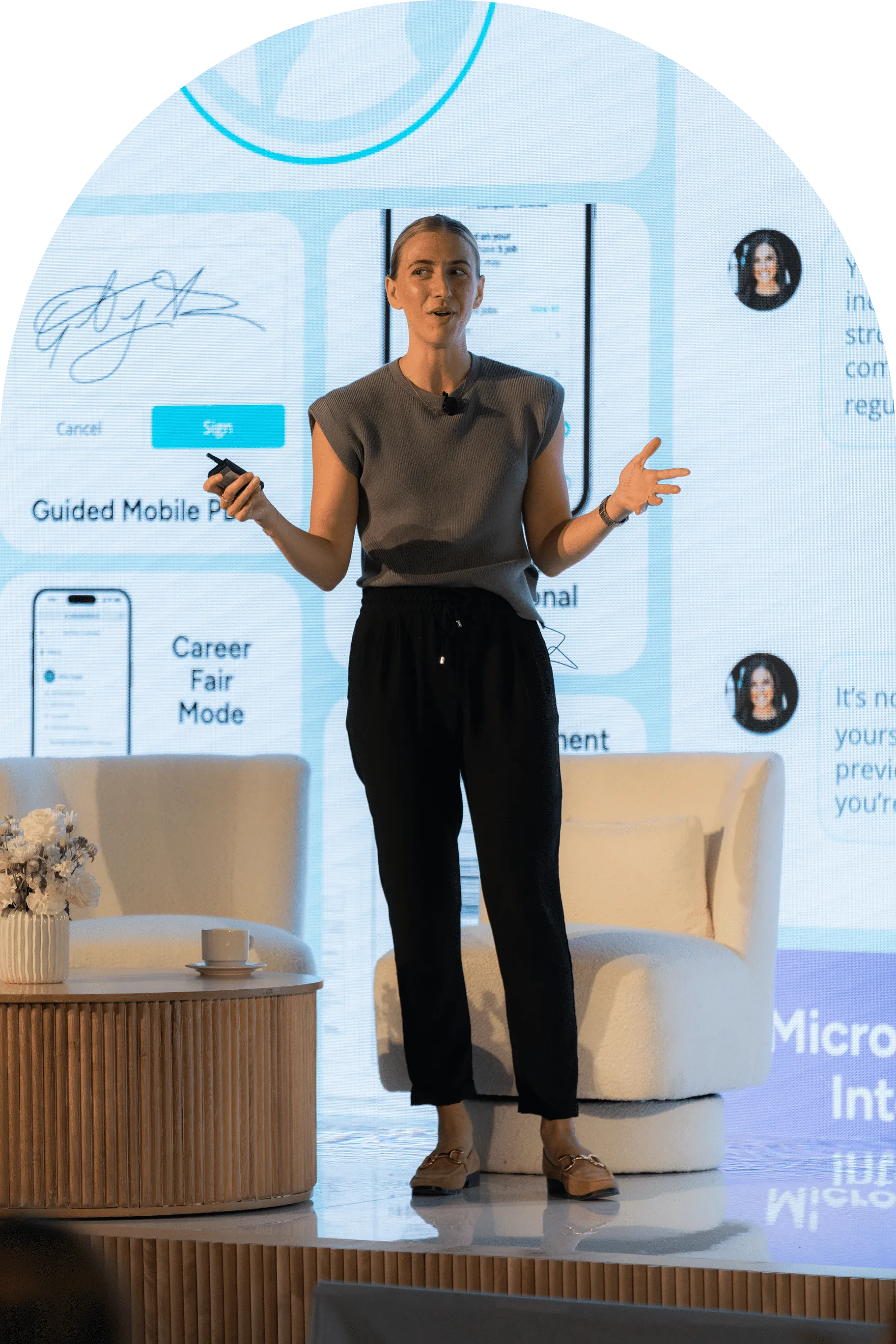 Woman in gray top and black pants giving a presentation on stage with digital screen behind her.