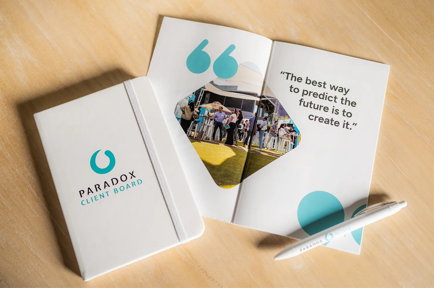 White Paradox Client Board notebook, open brochure with quote, and branded pen on a wooden table.