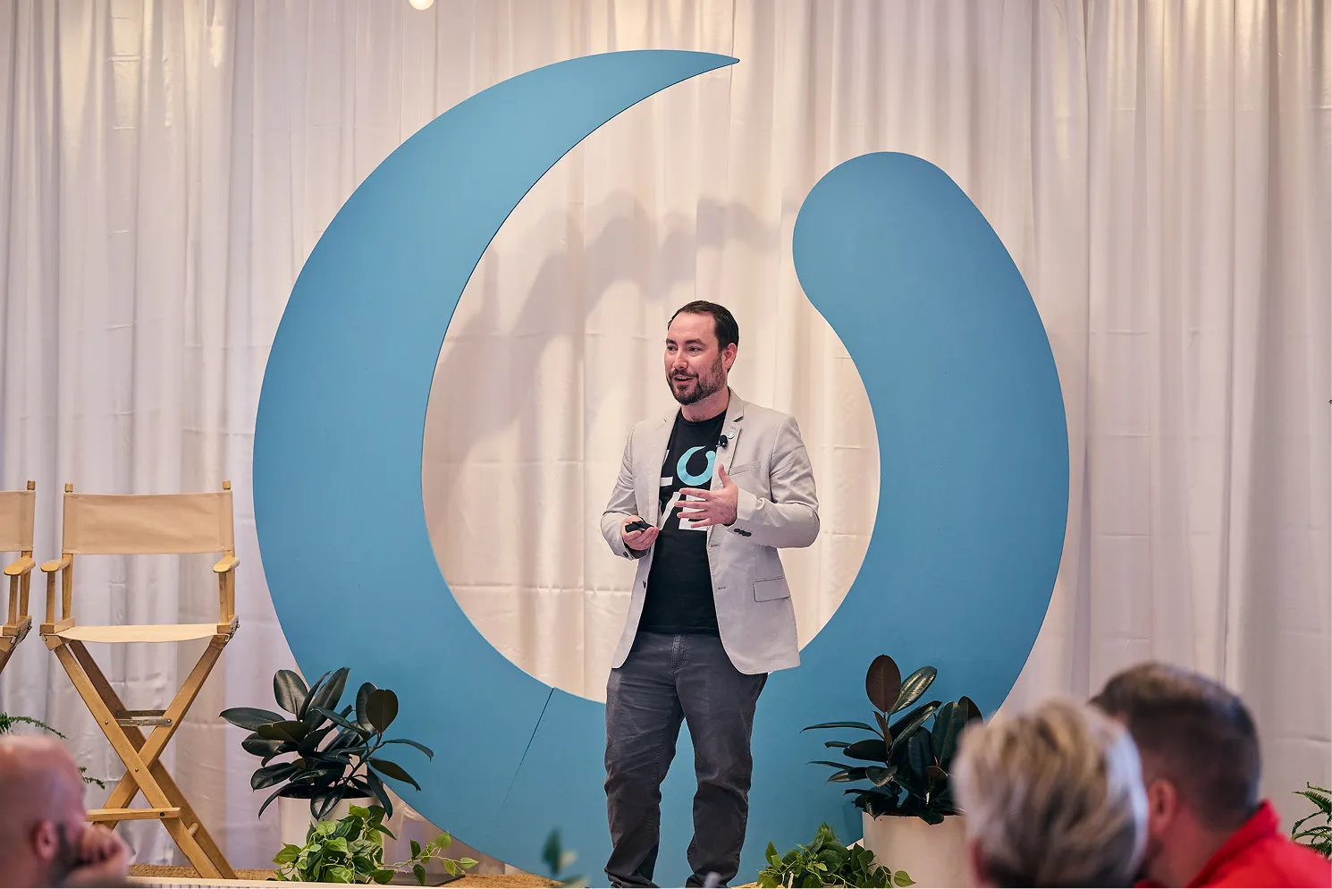 Man in gray blazer and black shirt speaking on stage in front of a large blue circular backdrop.