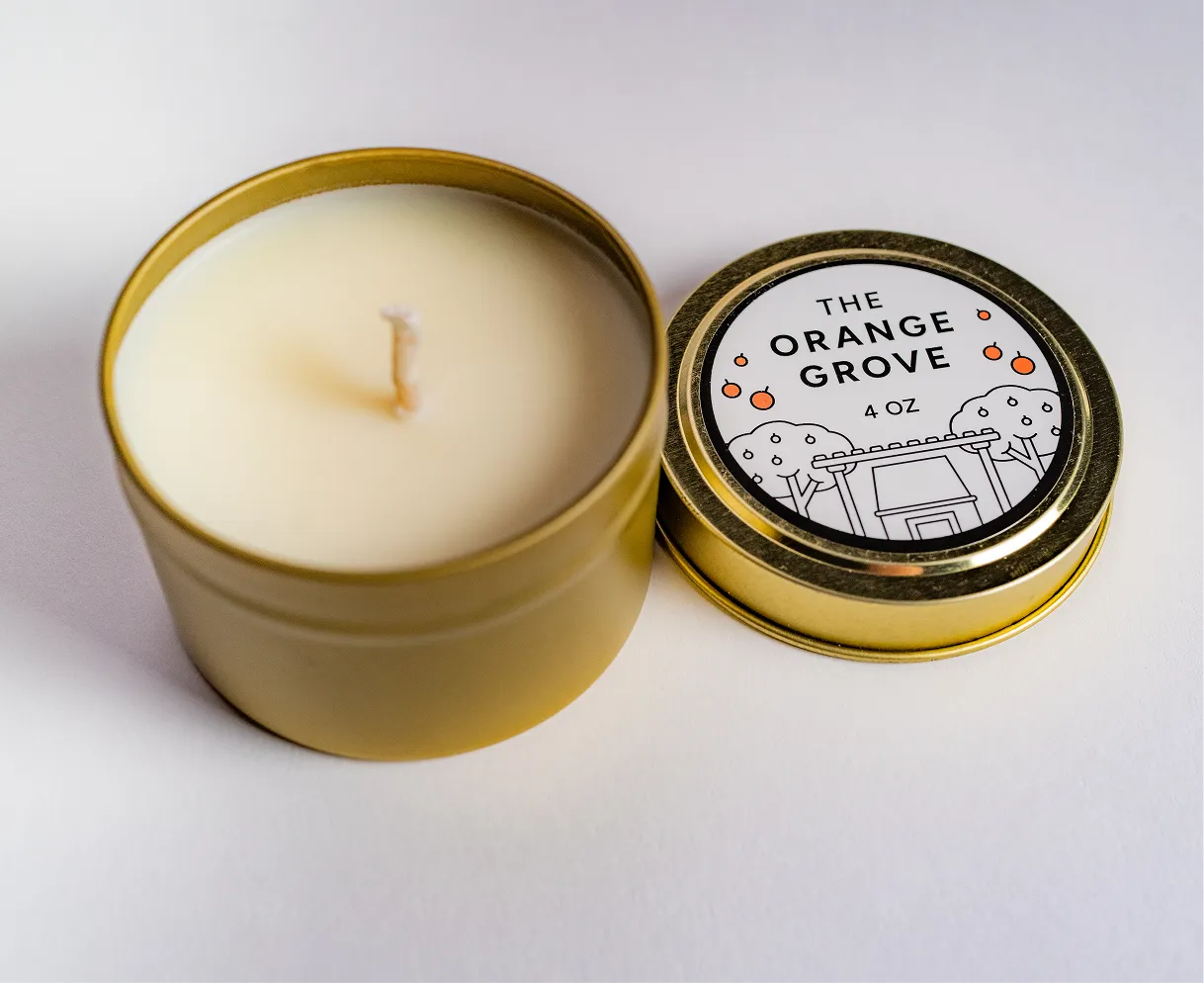 Open gold-colored tin candle with white wax and a lid labeled The Orange Grove 4 oz.