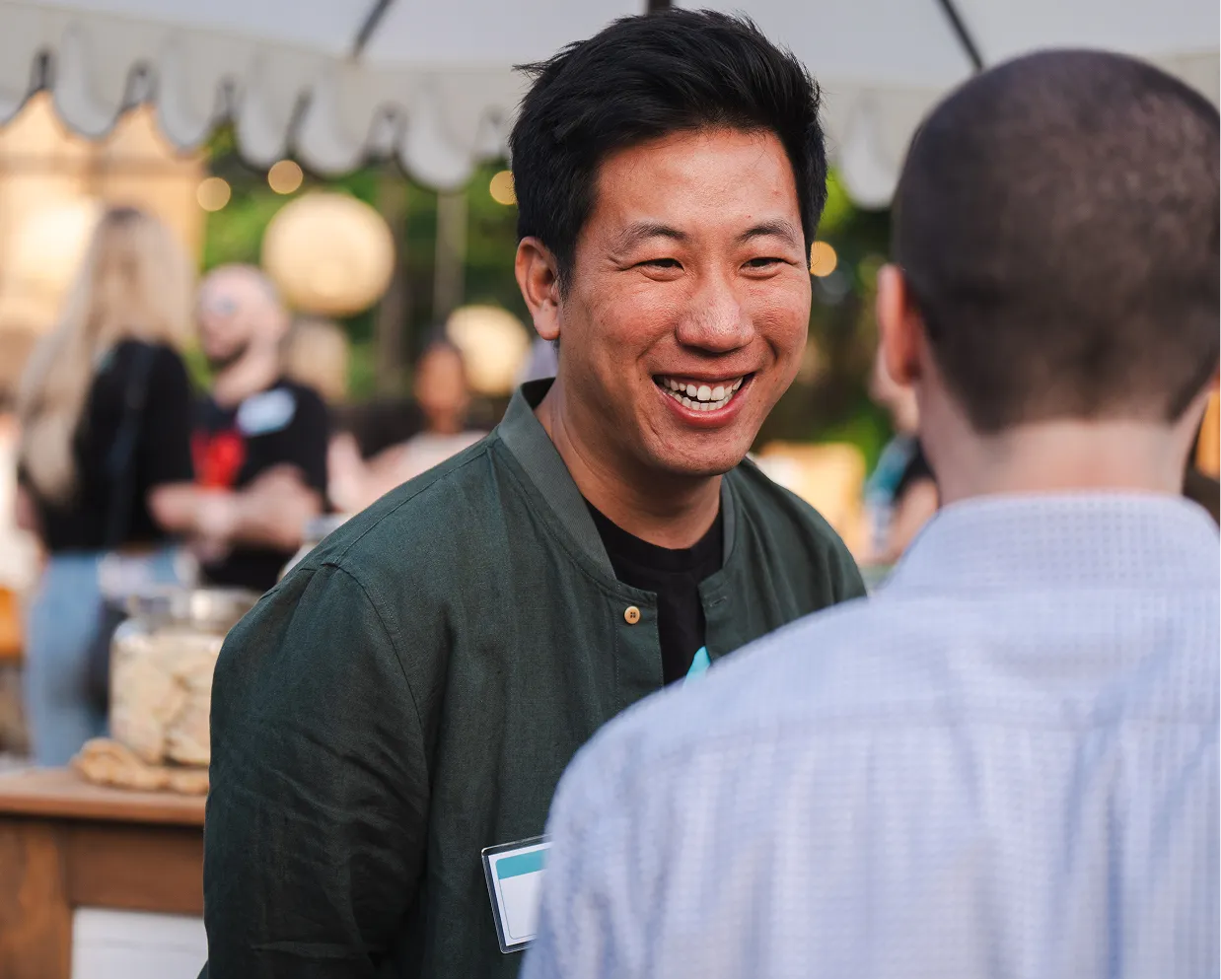 Smiling man in dark jacket talks with a man in a light patterned shirt at an outdoor event.