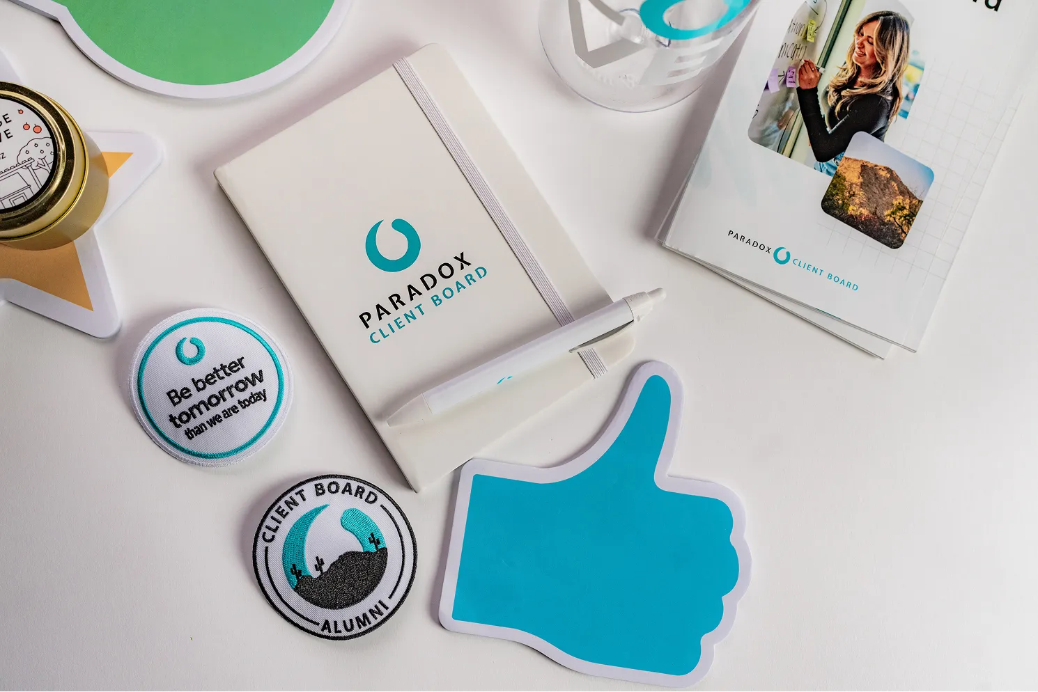 Paradox Client Board notebook, pen, patches, magazine, and blue thumbs-up cutout on a white surface.