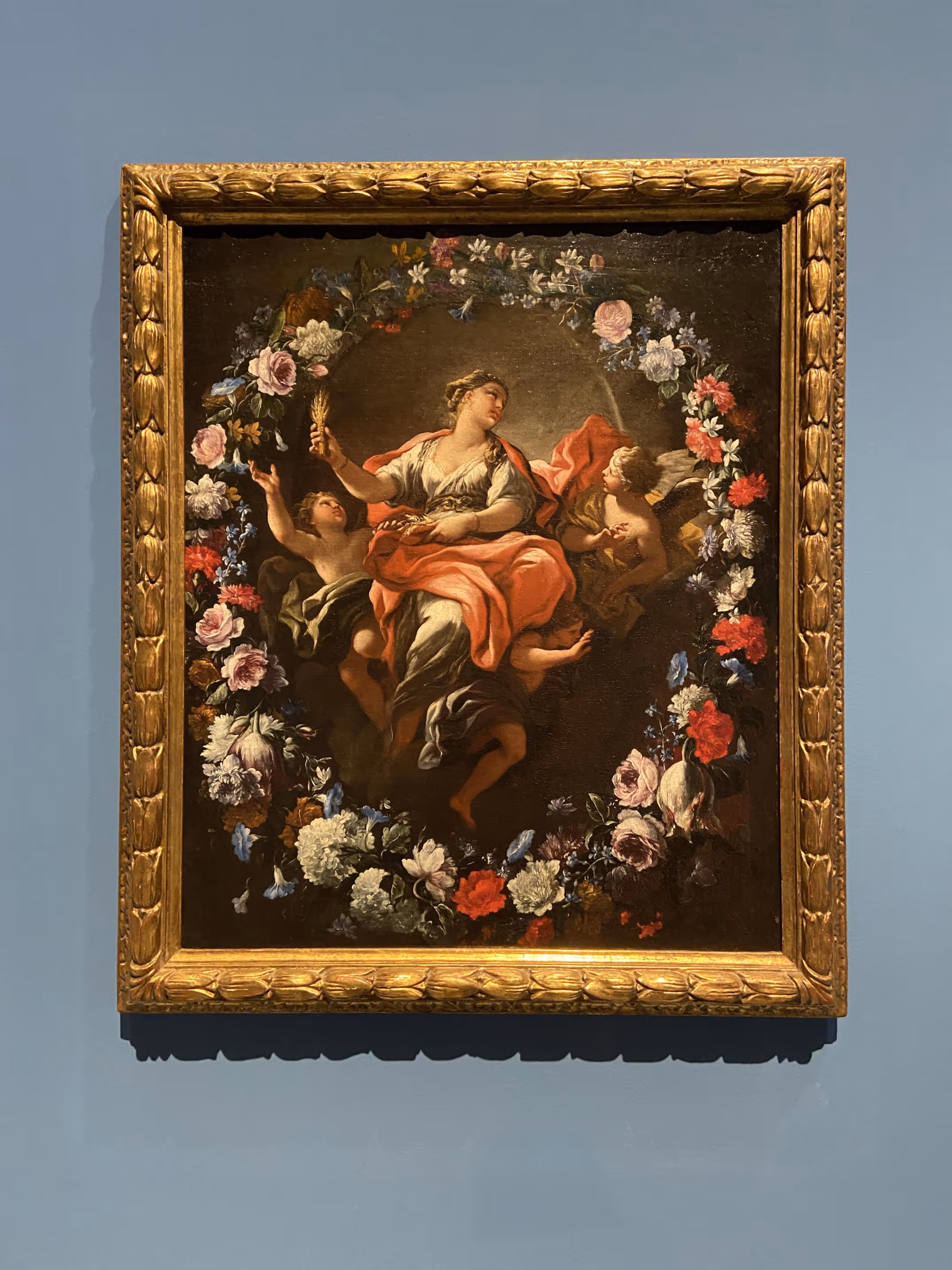 A gilt-framed classical oil painting of a reclining figure encircled by a garland of flowers and cherubs, hung on a grey-blue wall.