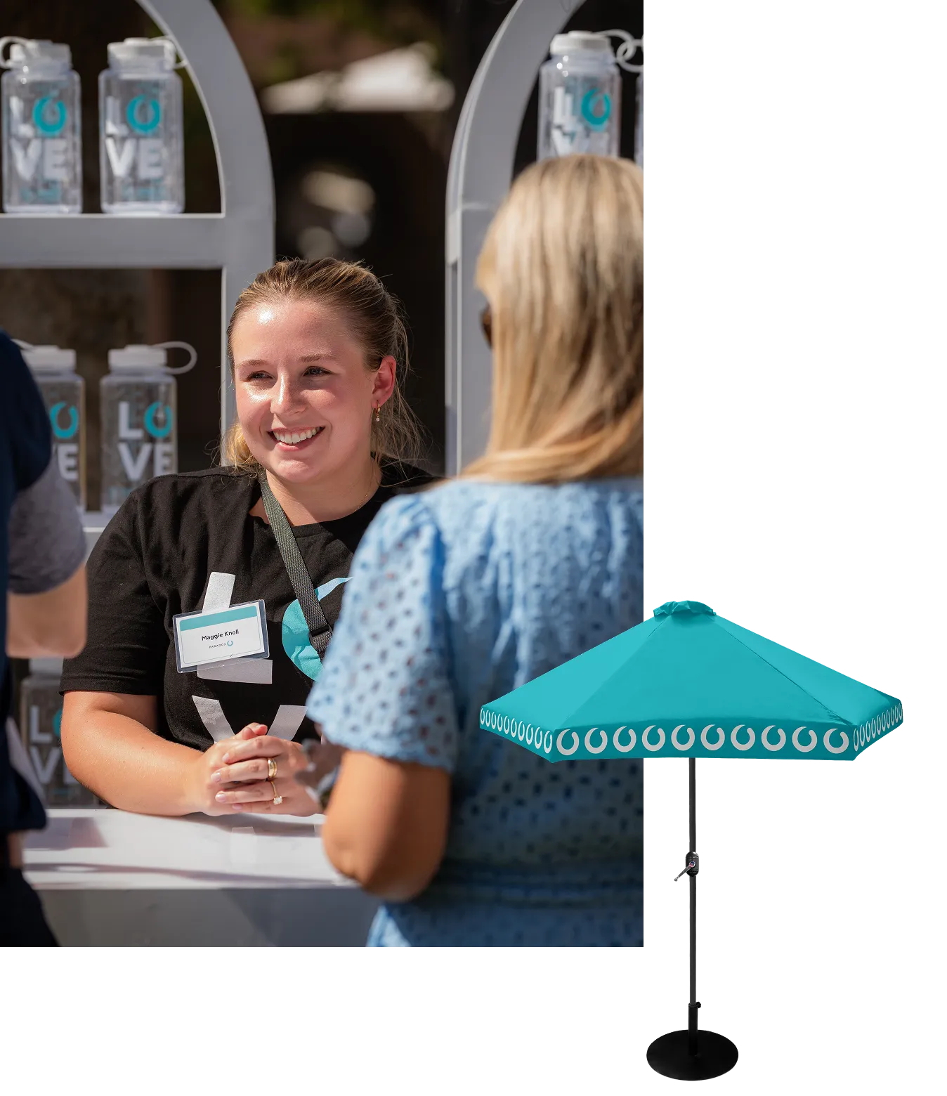 Smiling woman with name tag talks to a person in a blue dress at a booth with water bottles.
