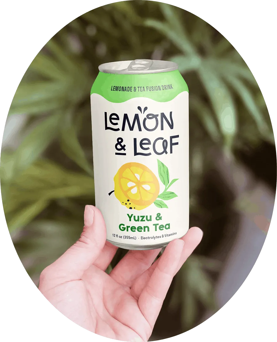 A hand holding a Lemon & Leaf Yuzu & Green Tea can in front of green foliage.