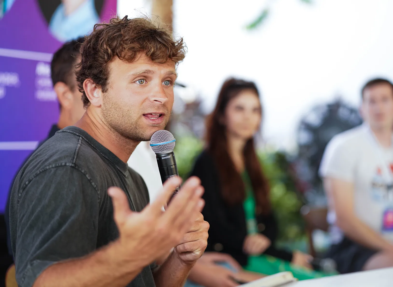 A man speaking into a microphone during an outdoor panel, with the audience behind him.