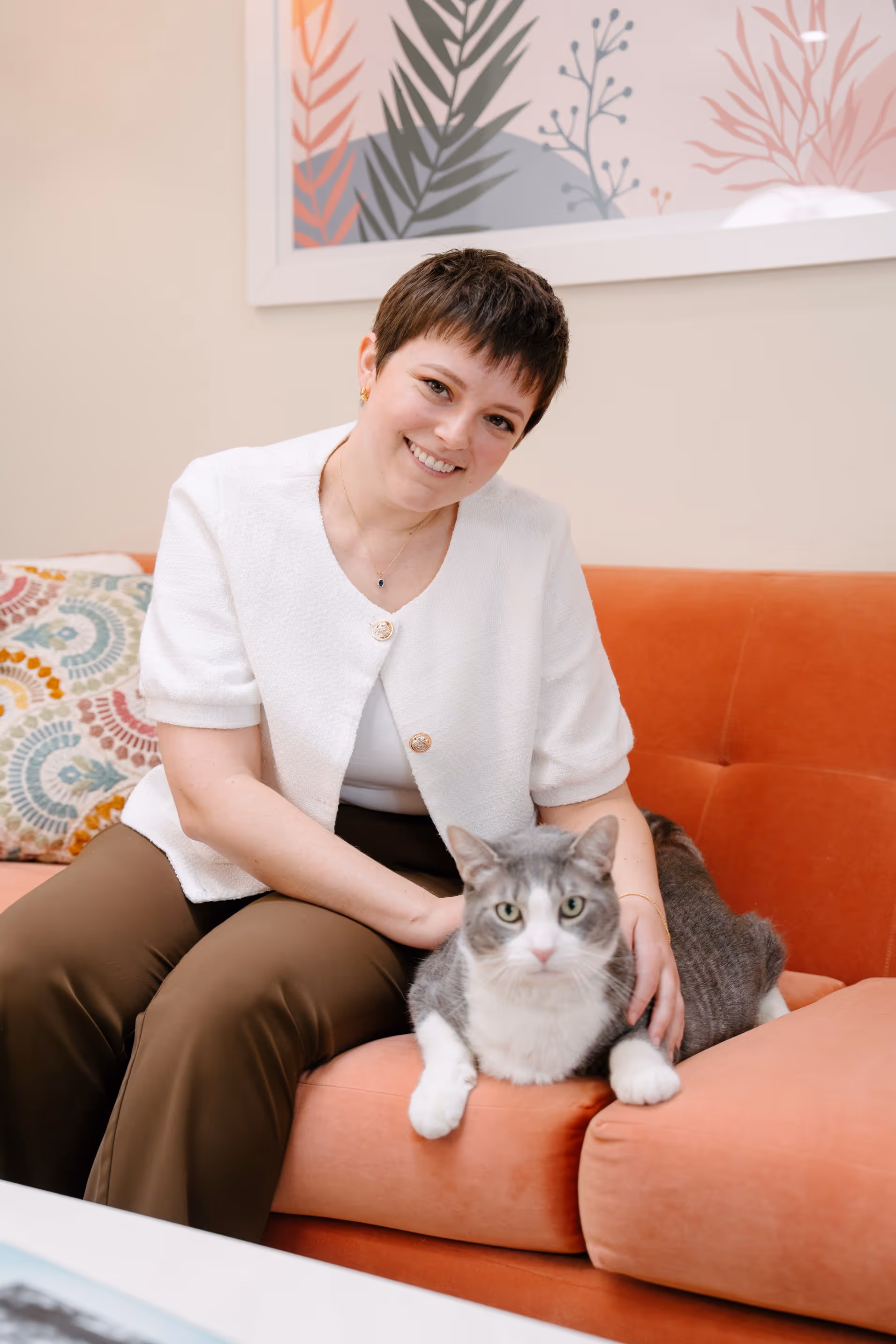 Maddie Moe seated on an orange sofa, smiling, with a grey and white cat resting on the sofa arm beside her.