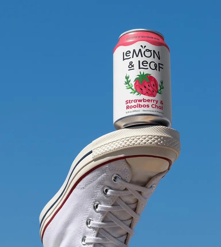 A Lemon & Leaf Strawberry can balanced on a white trainer against a blue sky.