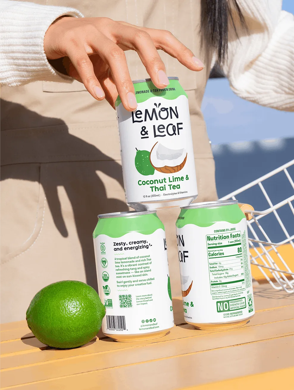 A hand reaching for Lemon & Leaf Coconut Lime & Thai Tea cans beside a fresh lime.