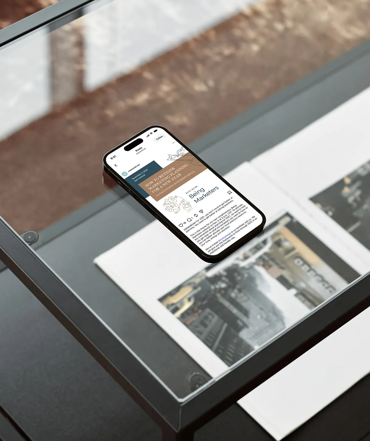 A phone on a glass table showing a Root + River article, beside a printed page.