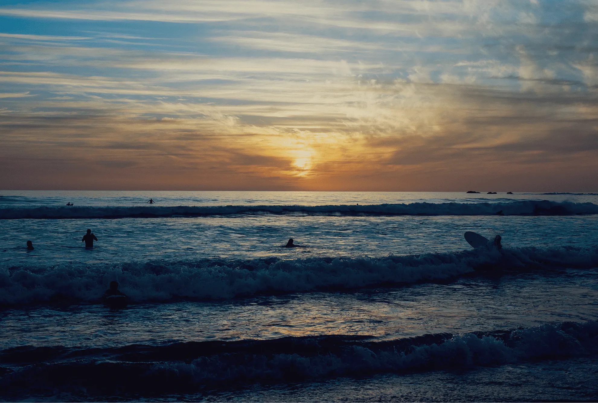 Silhouettes of surfers in ocean waves during a vibrant sunset with scattered clouds.
