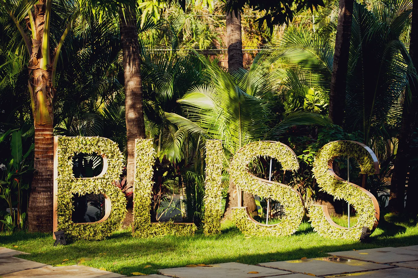 Large BLISS letters covered in green leaves on grass with tropical plants and palm trees behind.