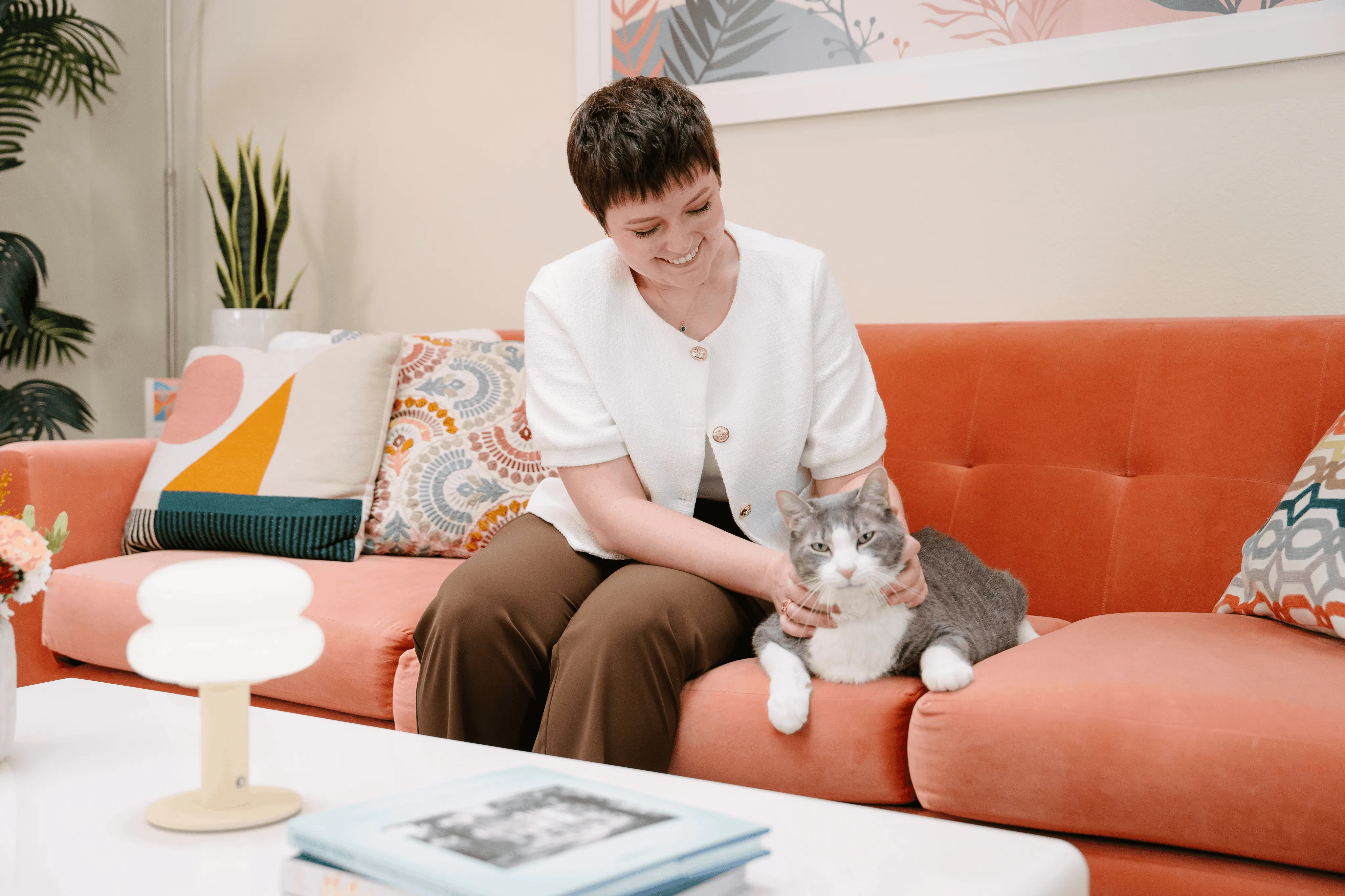 Maddie Moe sitting on an orange sofa, reaching towards a grey and white cat seated beside her.