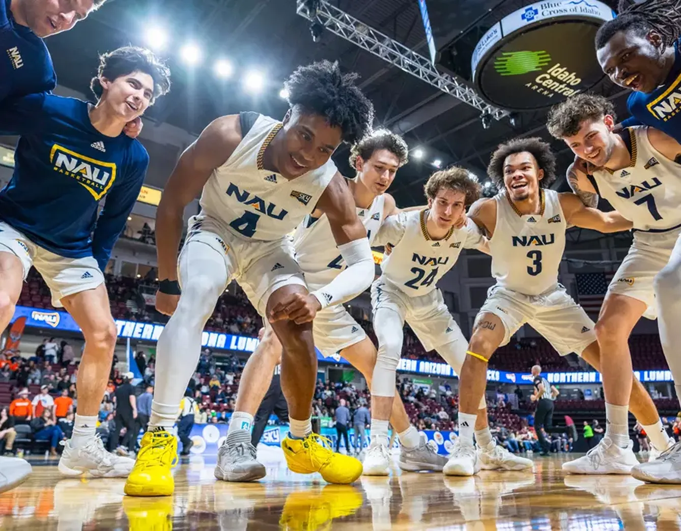 NAU basketball players in a huddle on the court, wearing white and blue uniforms in arena.
