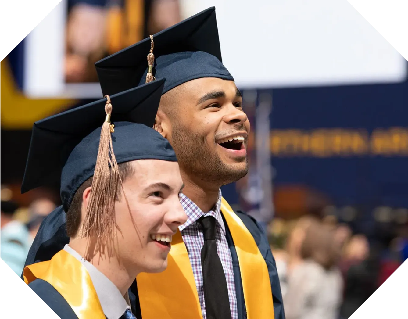 Two male graduates in caps and yellow stoles smiling and celebrating at a graduation ceremony.