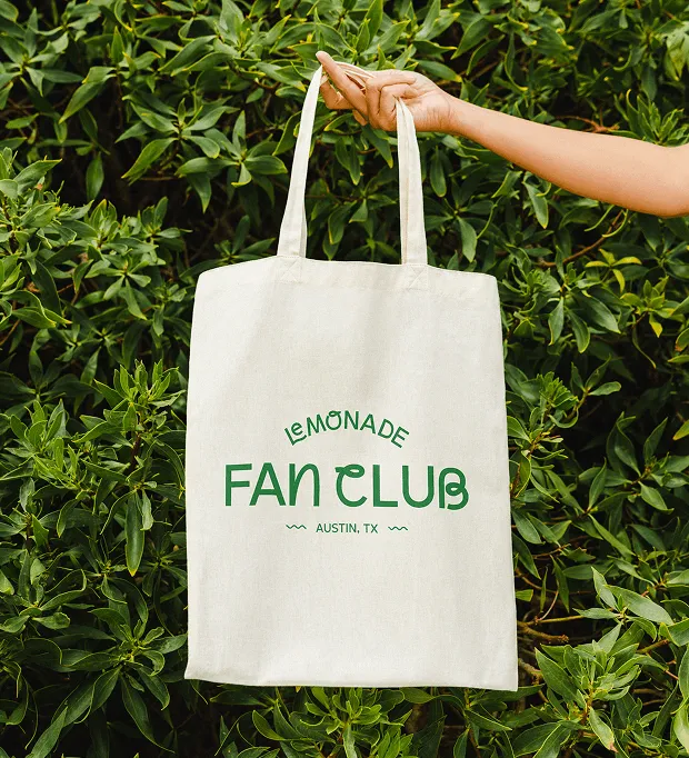 A hand holding a Lemonade Fan Club tote bag against green foliage.