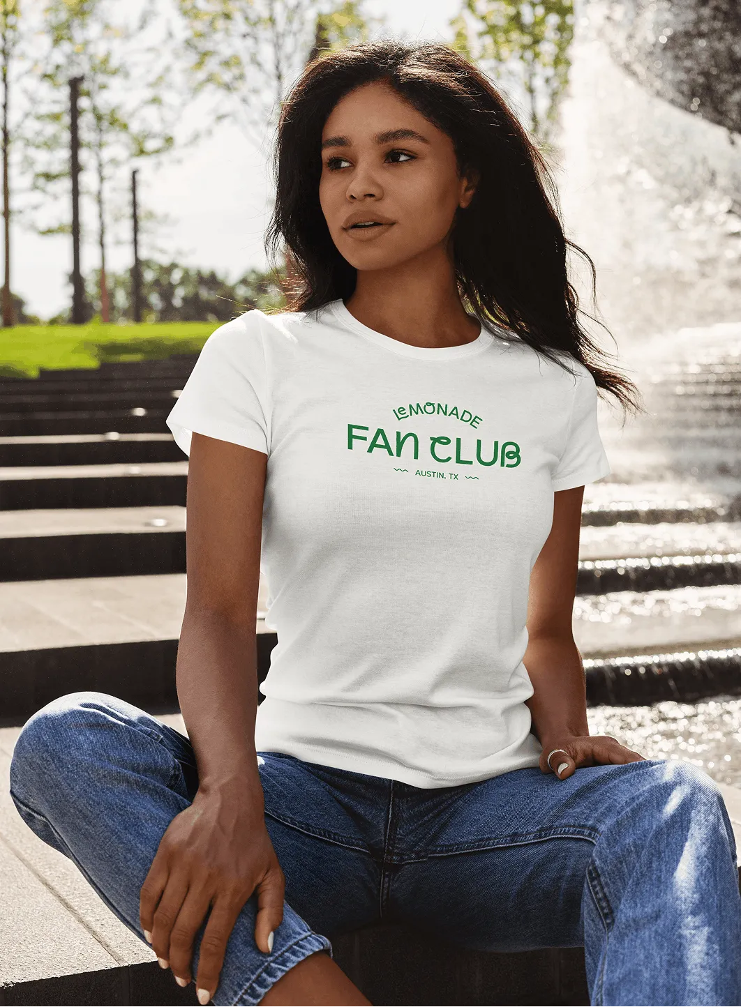 A woman sitting outdoors wearing a white Lemonade Fan Club t-shirt.