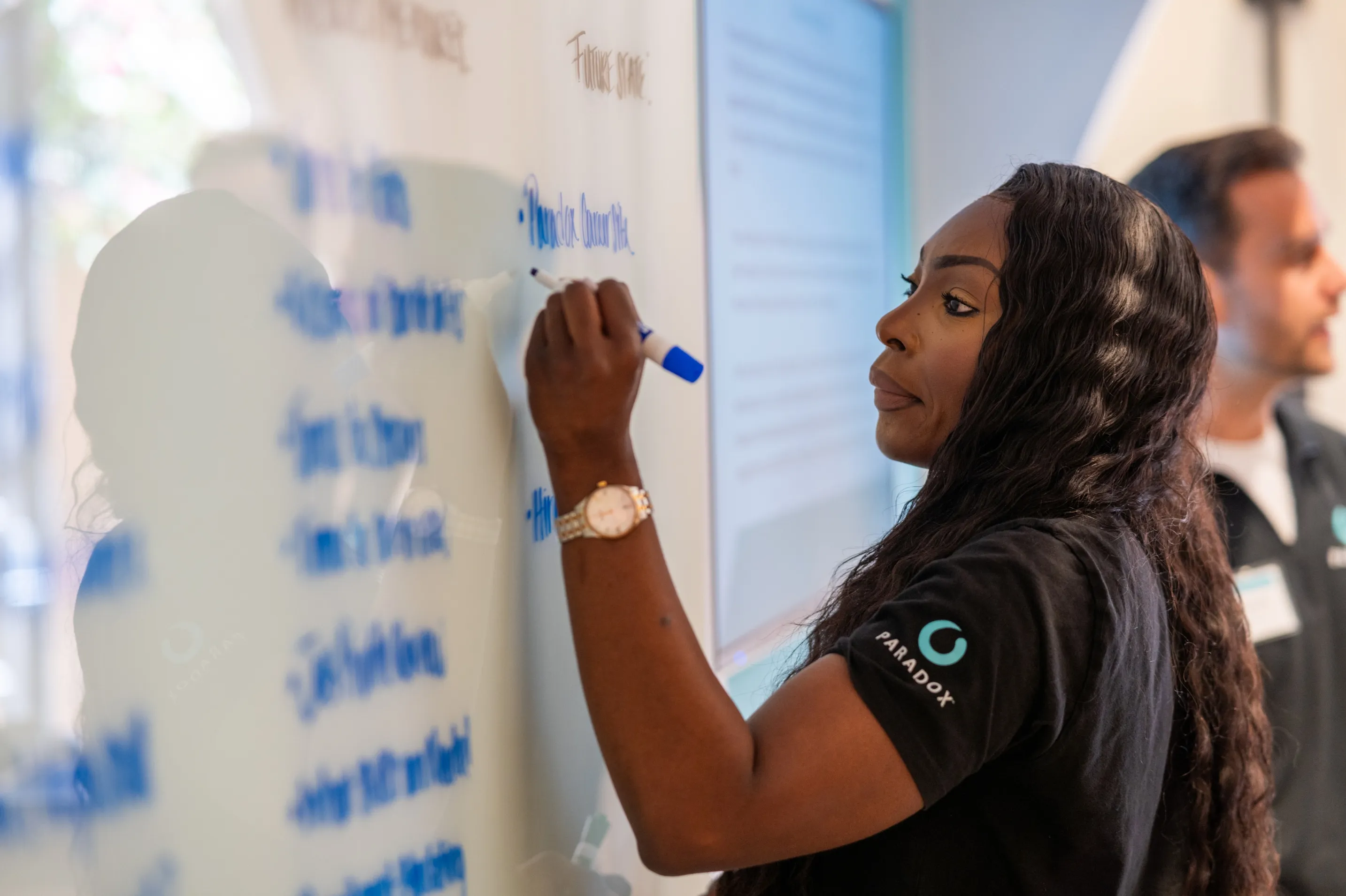 A woman in a Paradox t-shirt writing on a whiteboard during a working session.
