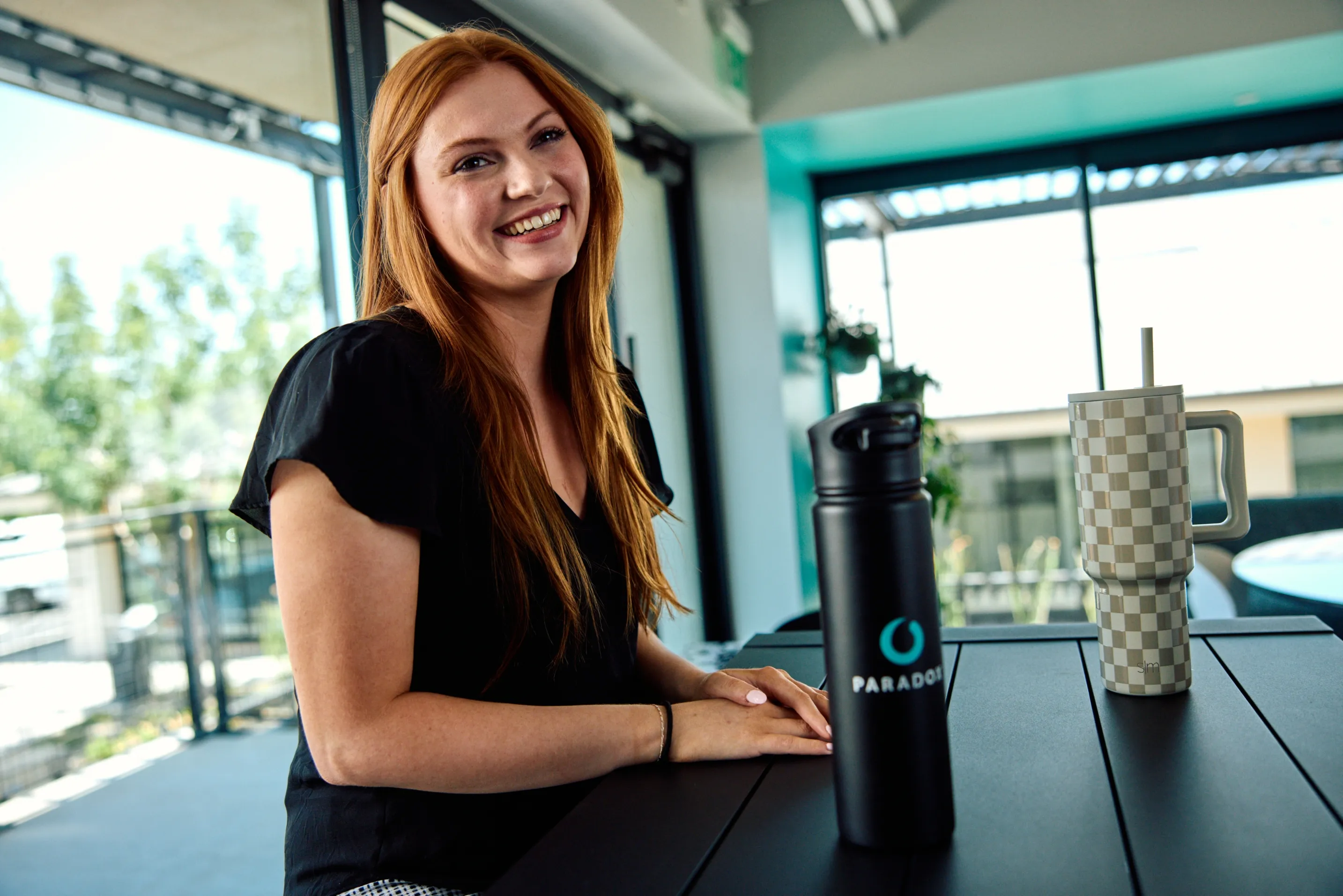 A woman smiling at a table beside a black Paradox water bottle and tumbler.