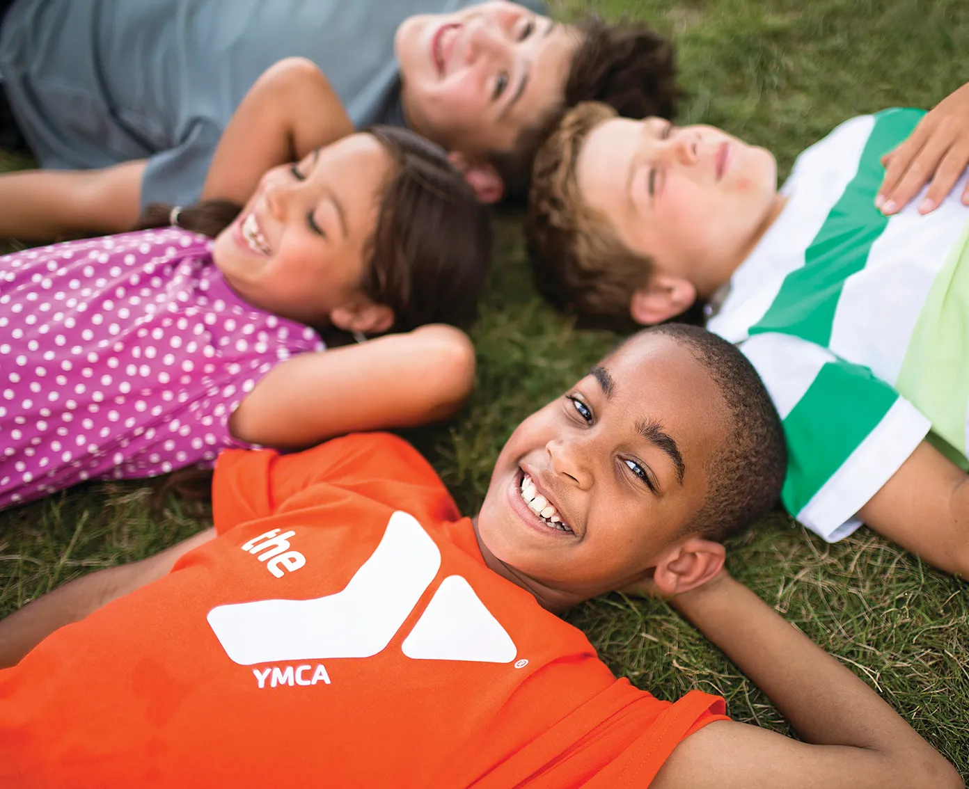 Children lying on grass in a circle, heads together, smiling upwards.