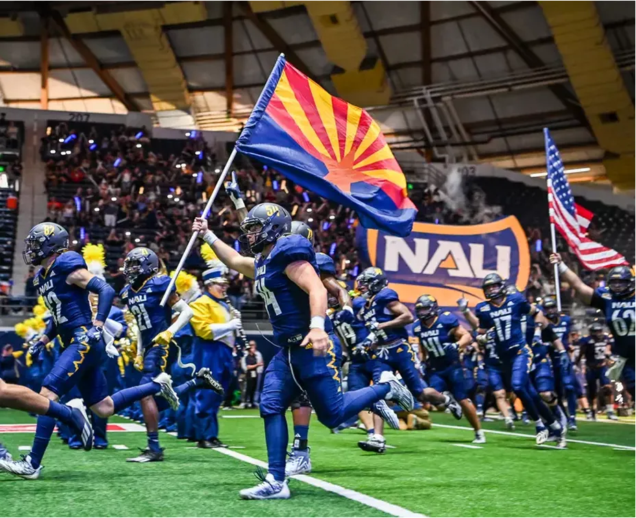 NAU football players running onto the field behind an Arizona flag and NAU banner.