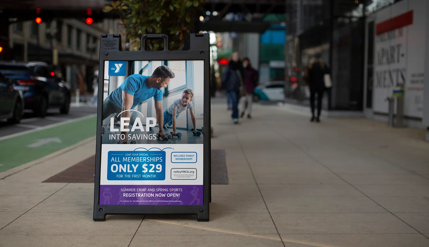 A YMCA "Leap into savings" A-frame sign on a pavement advertising $29 memberships.
