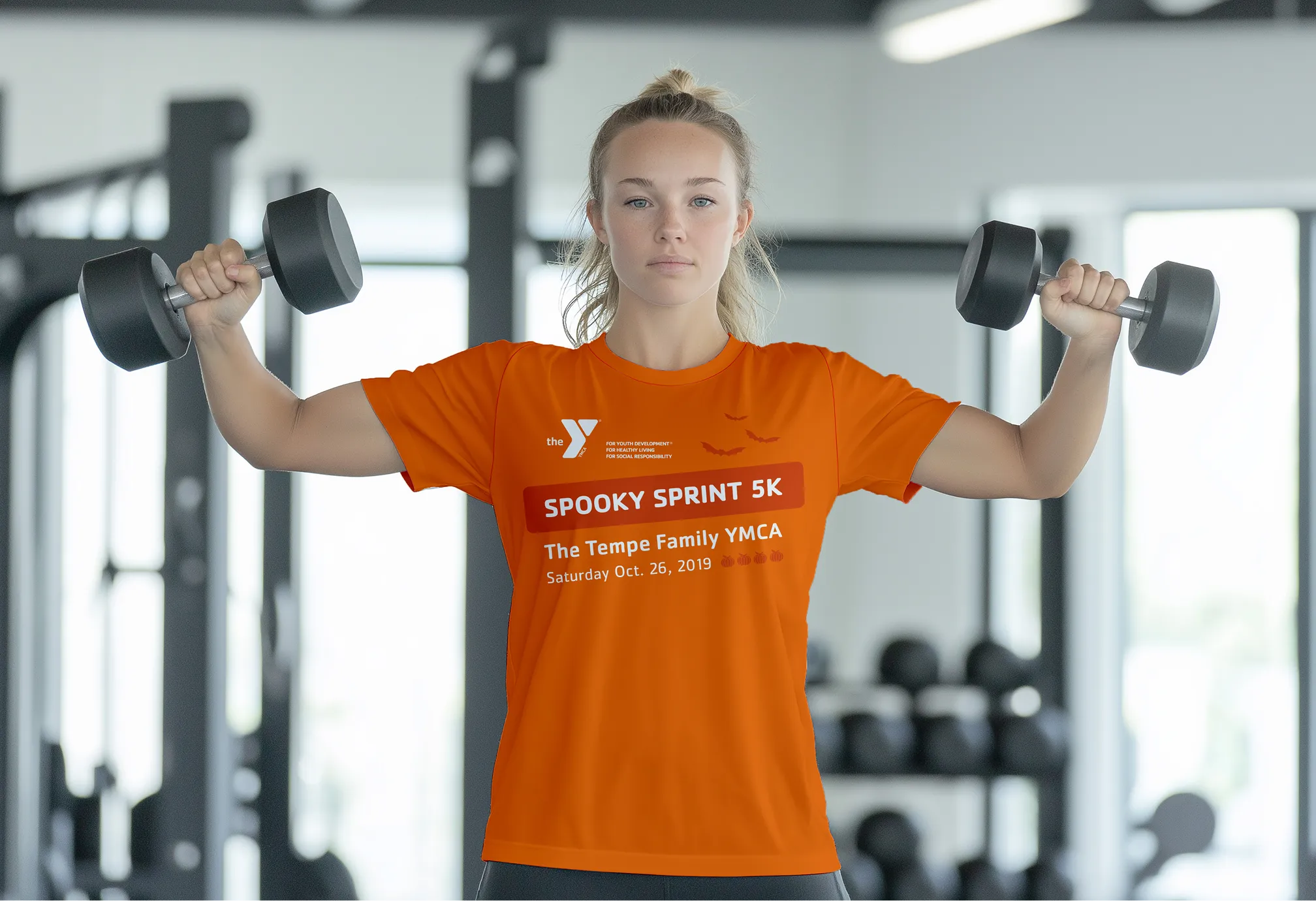 A woman in an orange Spooky Sprint 5K t-shirt lifting dumbbells in a gym.