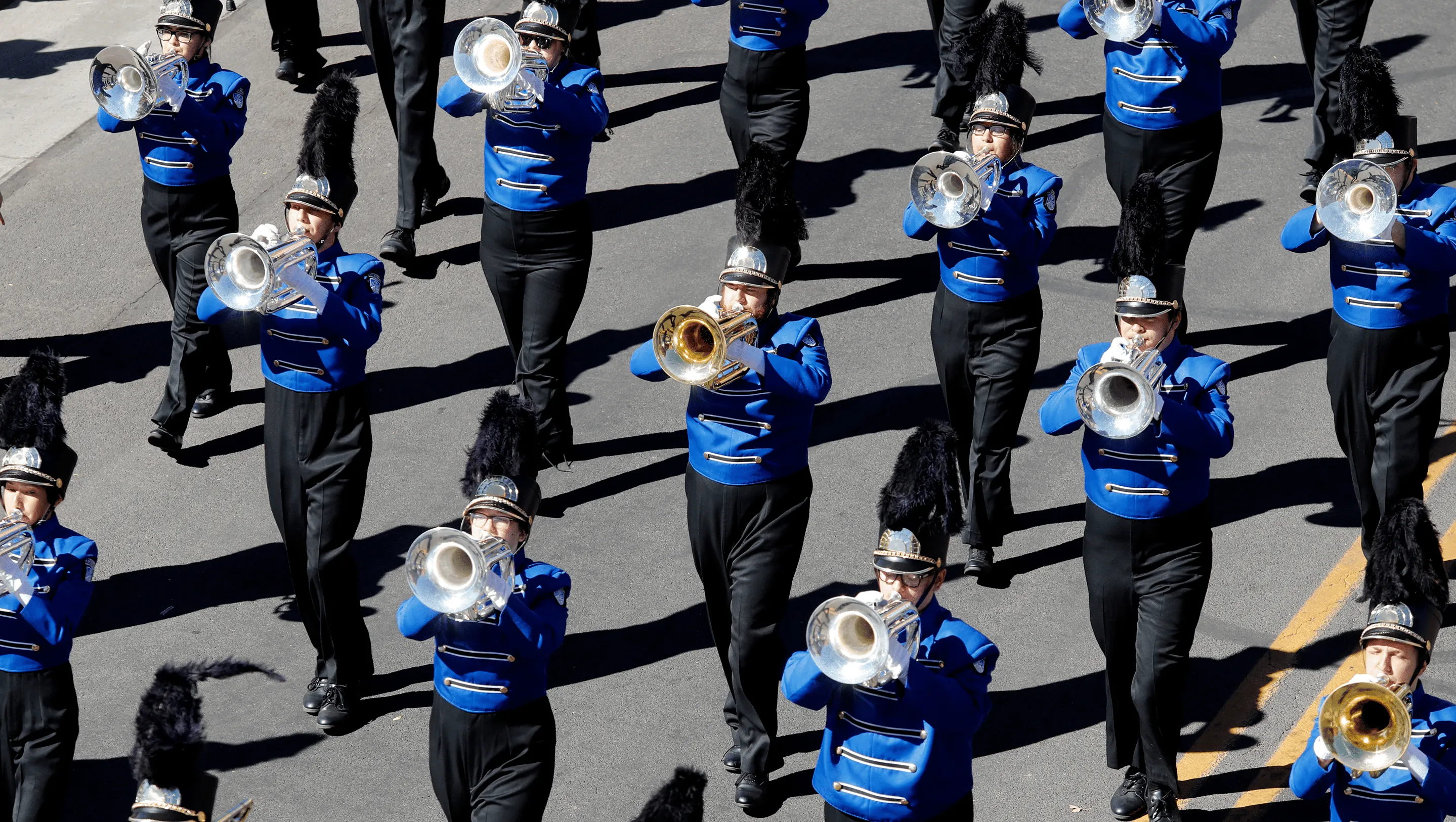 A marching band in blue uniforms and plumed hats playing brass in a parade.