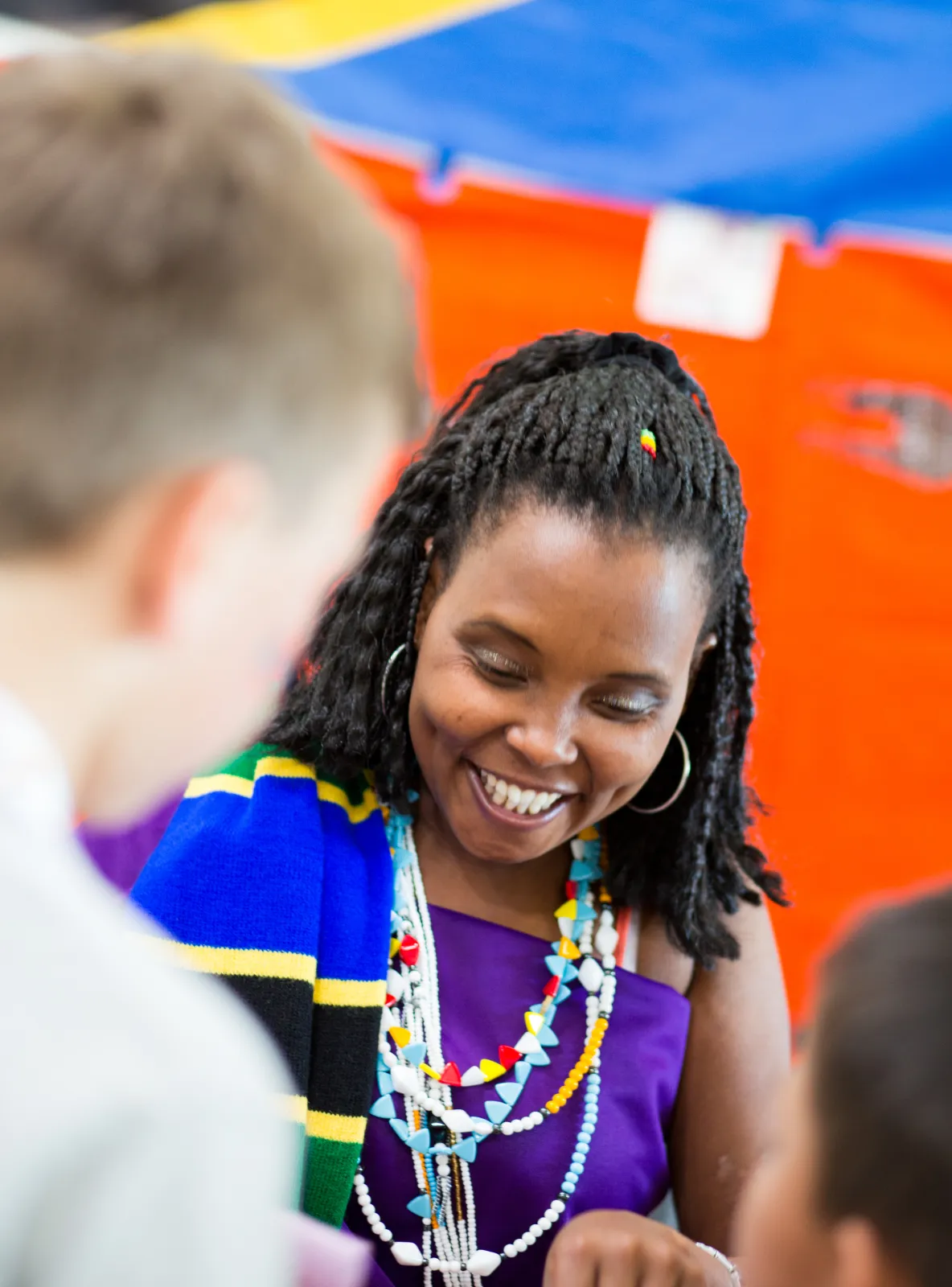 Smiling woman with braided hair wearing colorful beads and a blue, yellow, green shawl.