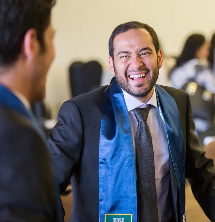 Man in graduation gown and tie laughing and talking with another person at an indoor event.