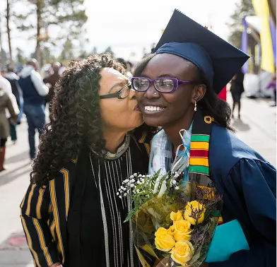 Woman kisses young graduate in cap and gown holding bouquet of yellow flowers outdoors.