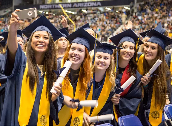 Group of five female graduates in caps and gowns smiling, holding diplomas, and taking a selfie.