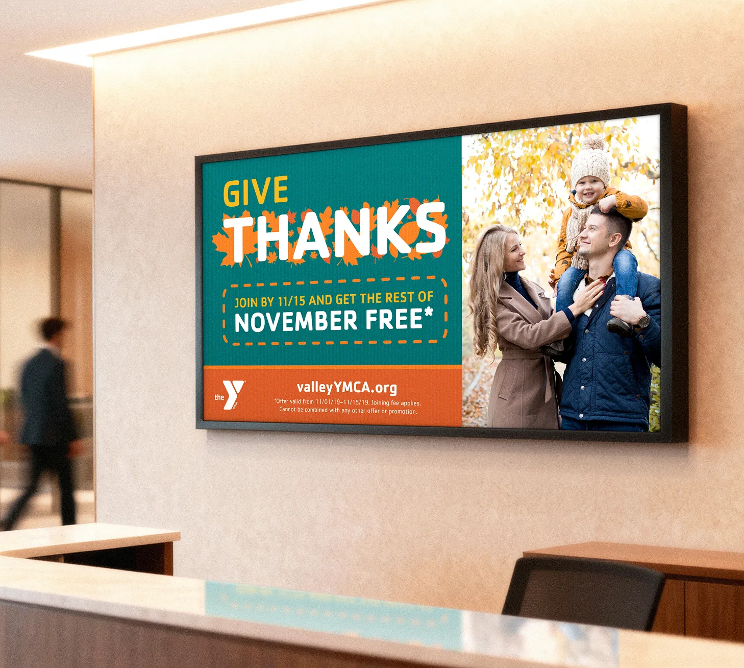 A wall-mounted screen in a lobby showing a YMCA "Give Thanks" November free campaign.