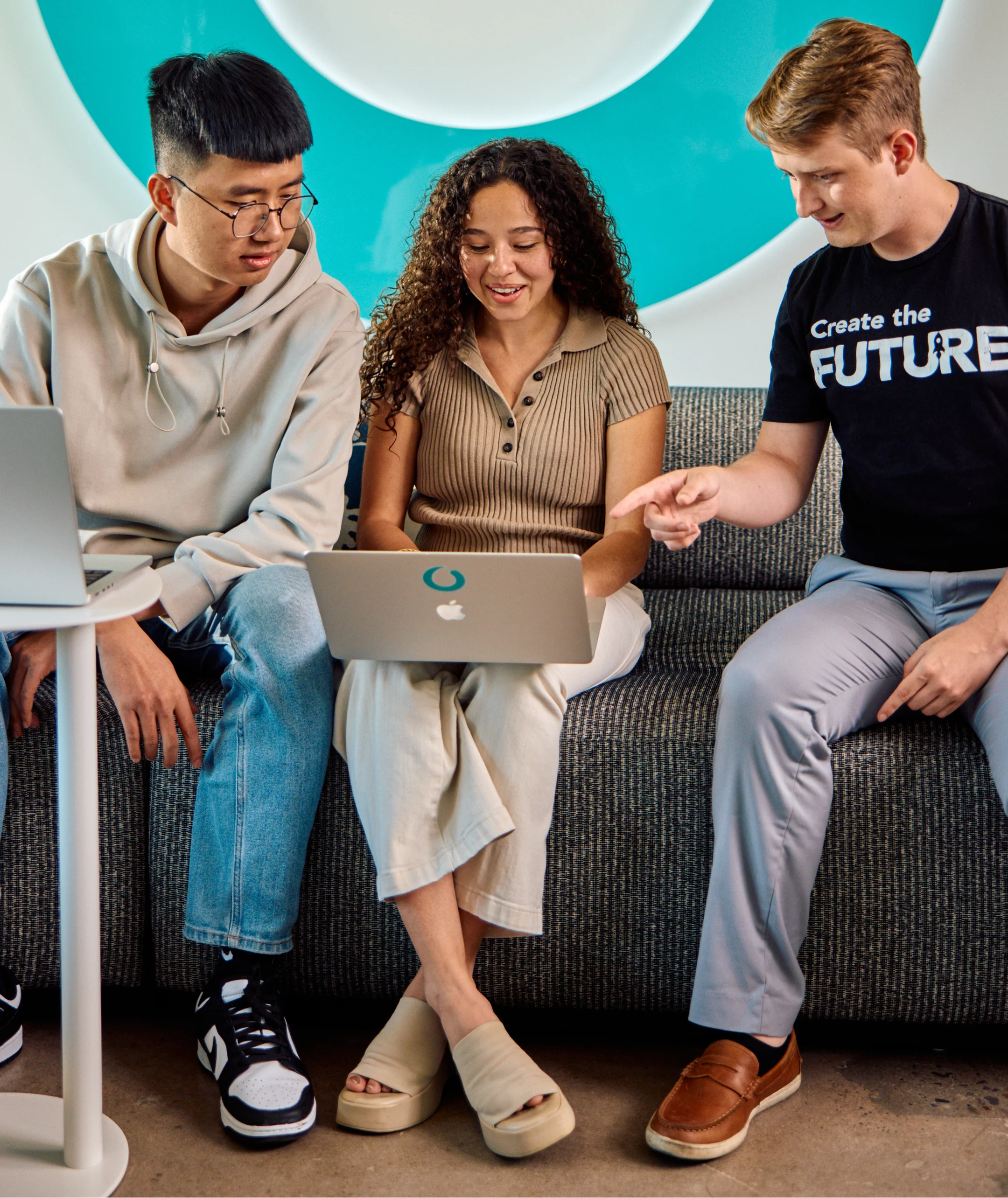 Three colleagues on a bench against a teal wall, one in a Create the Future t-shirt, using laptops.