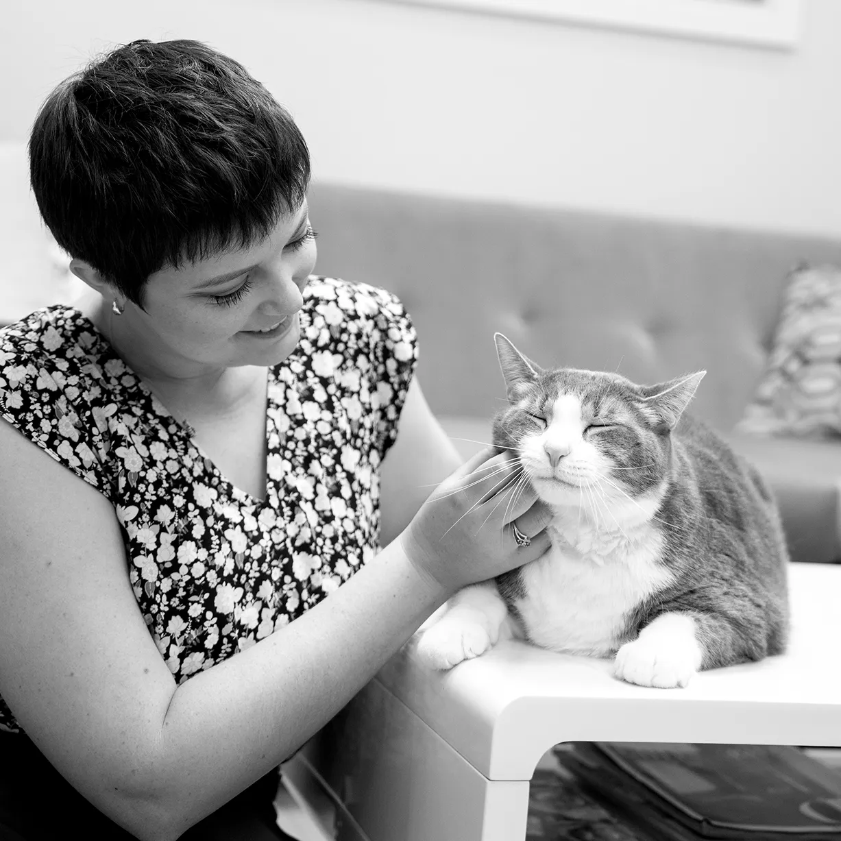 A black and white photograph of Maddie Moe smiling as she strokes a grey and white cat lying on a white surface.