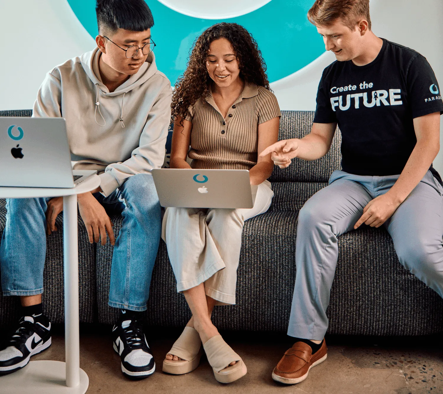 Three colleagues seated on a sofa with laptops, talking and laughing together.