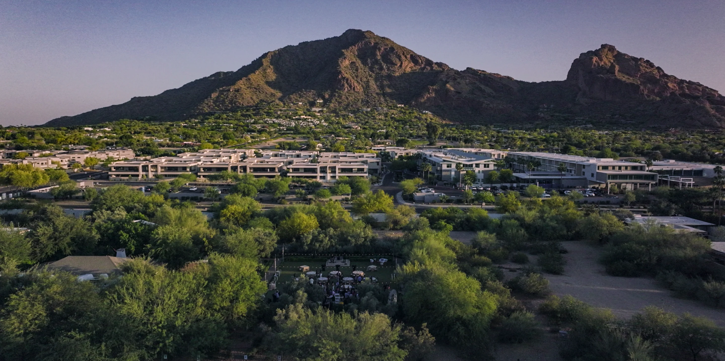 Residential area with green trees and cars, set against large rocky mountains under a clear sky.