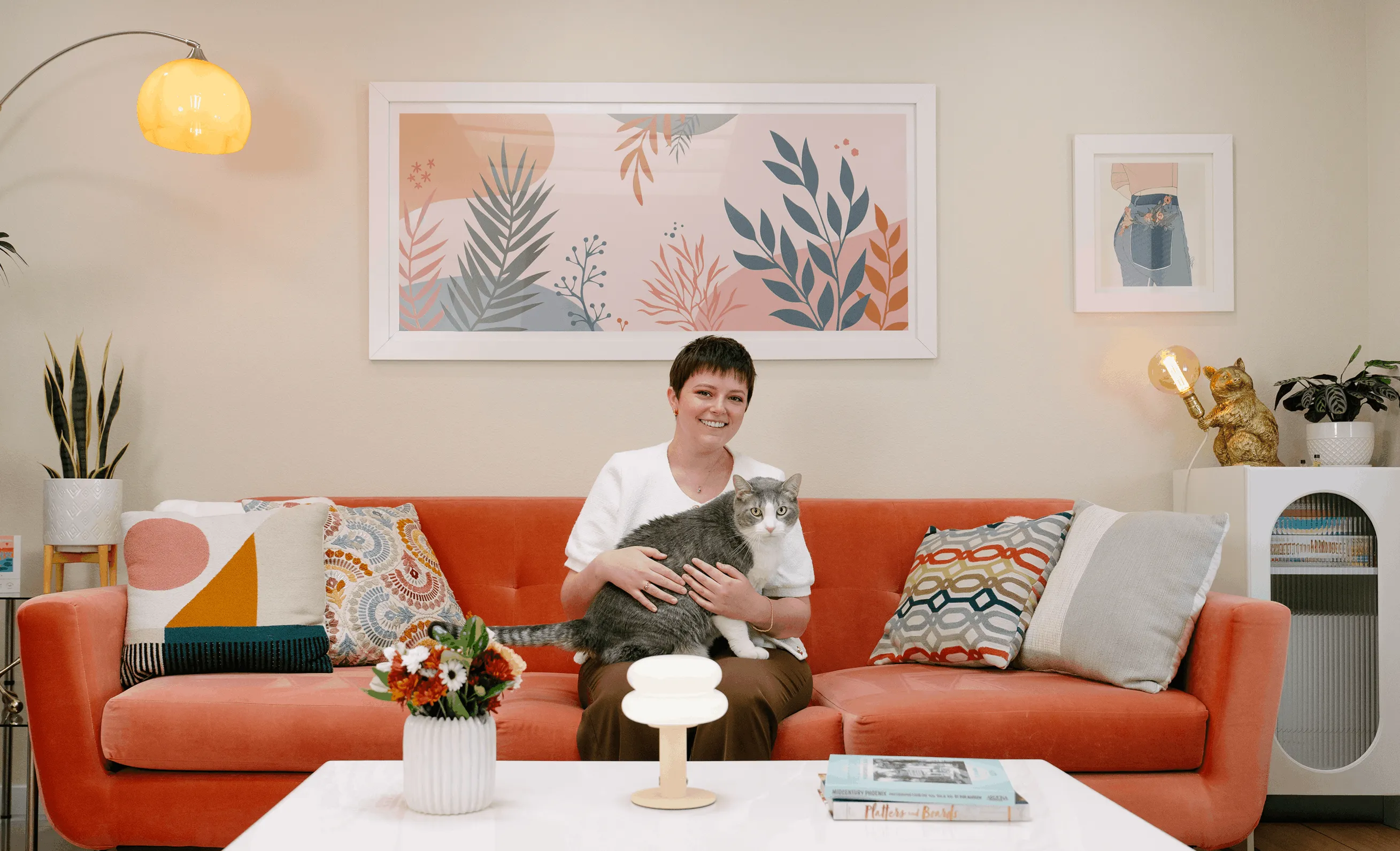 Maddie Moe sitting on a bright orange sofa in a sunlit living room, holding a grey and white cat, with framed botanical artwork on the wall behind her.