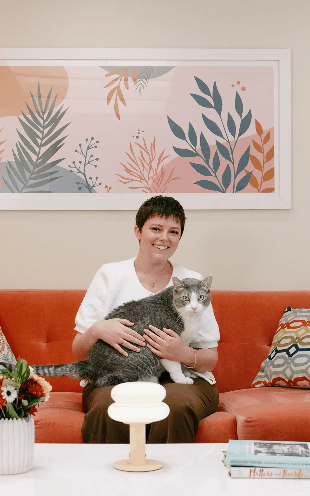Maddie Moe sitting on a bright orange sofa in a sunlit living room, holding a grey and white cat, with framed botanical artwork on the wall behind her.