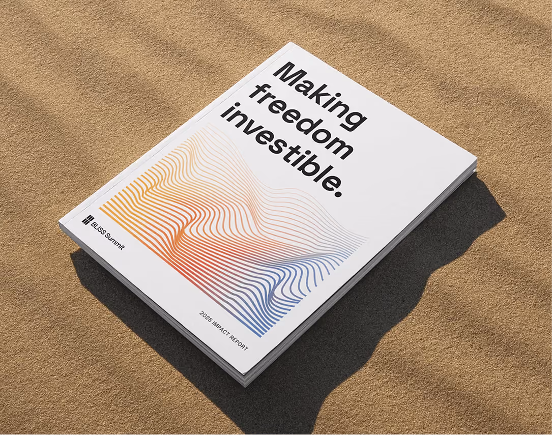 A printed impact report lying on rippled sand, its cover reading "Making freedom investible." above a rainbow contour-line illustration.