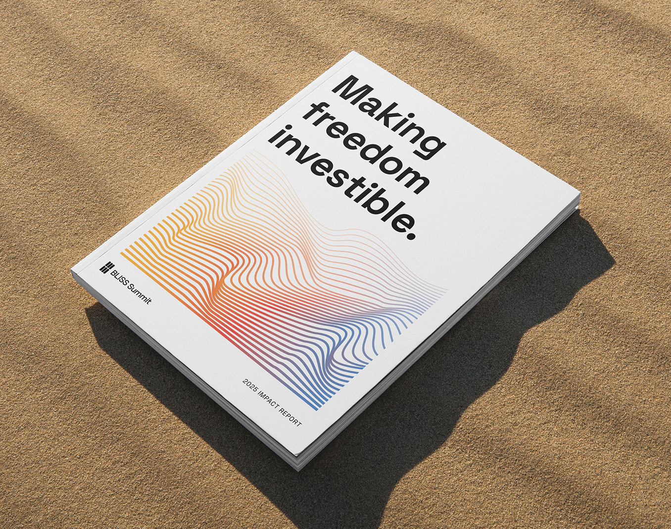 A printed impact report lying on rippled sand, its cover reading "Making freedom investible." above a rainbow contour-line illustration.