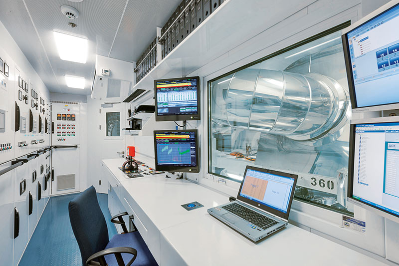Modern control room with multiple monitors, a laptop, control panels, and a large window showing industrial equipment outside.