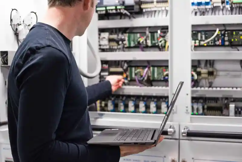 Technician holding a laptop and working on an industrial control panel with wiring and circuit boards.