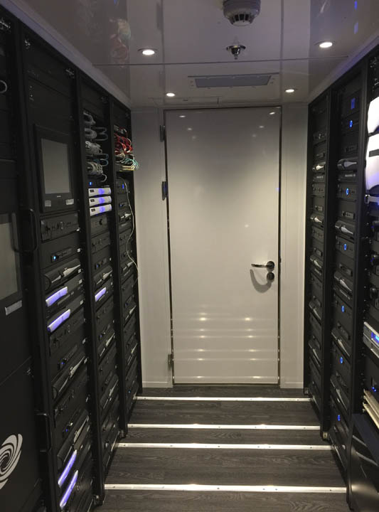 Narrow room with server racks lining both walls and a closed white door at the end, with recessed ceiling lights and dark floor panels.