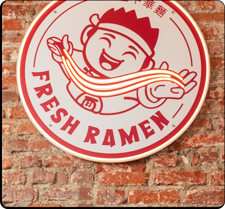 Round neon sign with a happy chef holding noodles and the words Fresh Ramen on a brick wall.