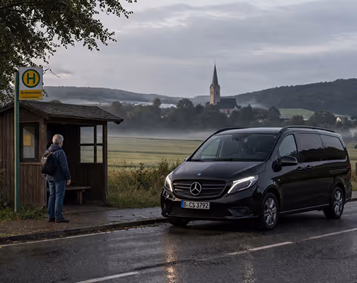 Eine Person wartet an einer Bushaltestelle auf einem ländlichen Weg, daneben steht ein schwarzer Mercedes-Van.