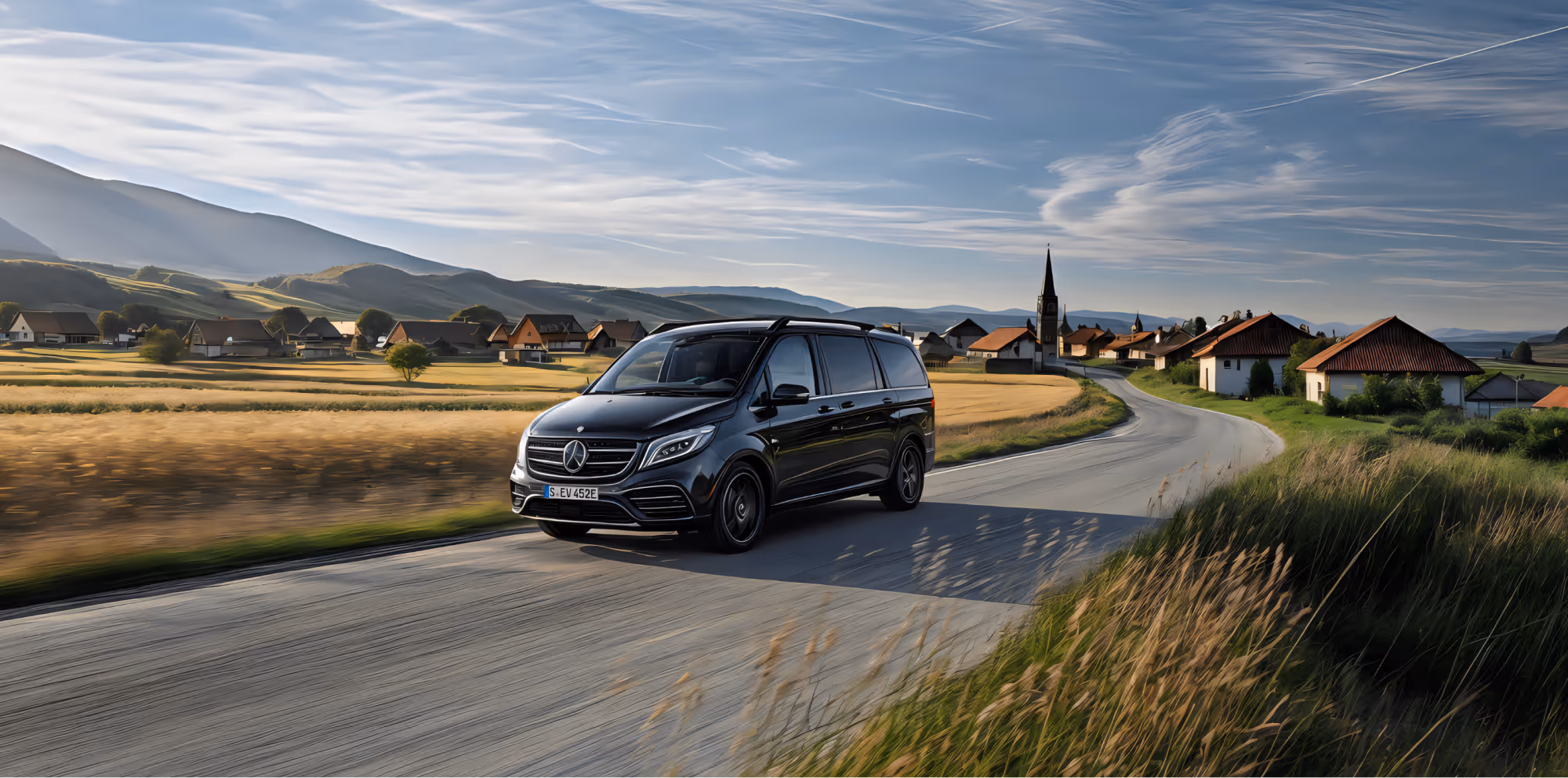 Schwarzer Mercedes-Benz Van fährt auf Landstraße mit Feldern und Dorf im Hintergrund.