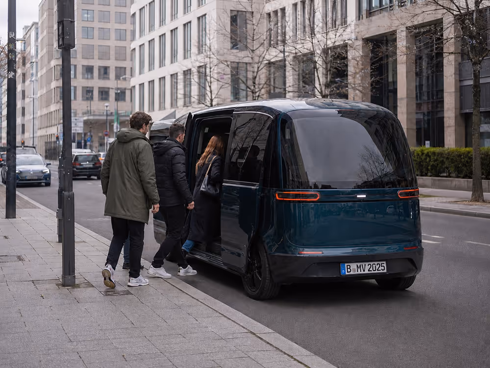 Drei Personen steigen in einen modernen, schwarzen Minivan an einer städtischen Straße ein.