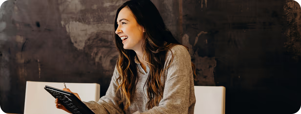 Smiling woman with long hair holding a tablet and stylus seated against a dark textured wall.