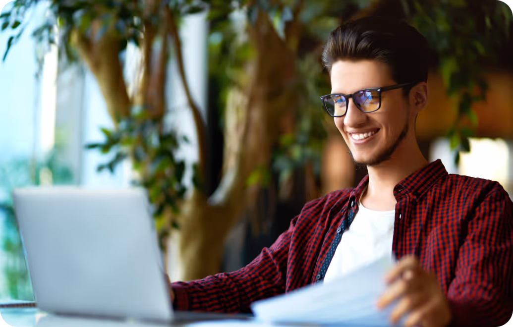 Smiling young man in glasses working on a laptop while holding papers in a green, indoor setting.